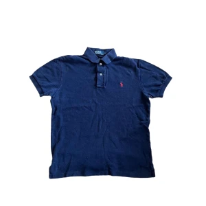 Ralph Lauren piké  - Klassisk marinblå pikétröja från Ralph Lauren med korta ärmar och krage. Tröjan har knappar framtill och den ikoniska röda loggan broderad på bröstet. Tillverkad i mjuk bomull som känns skön mot huden. Perfekt för en clean och sportig stil.  Jag skickar ej bild när jag bär plaggen och jag vet ej vilken längd och vikt de passar då jag inte prövar kläderna jag säljer 😃 Lite blekt färg annars i bra skick !