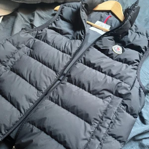 Svart dunväst från Moncler - Säljer en svart dunväst från Moncler med klassisk quiltad design och hög krage. Västen har dragkedja framtill och Moncler-logga på bröstet. Kvitto finns, skriv för frågor video på när den köps finns 