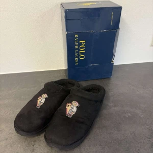 Svarta tofflor Polo Ralph Lauren - Mysiga svarta tofflor från Polo Ralph Lauren med broderad nalle i vinteroutfit framtill. Utsidan är i mjuk mocka och insidan är fodrad för extra komfort. Perfekta att glida runt i hemma när det är kallt.