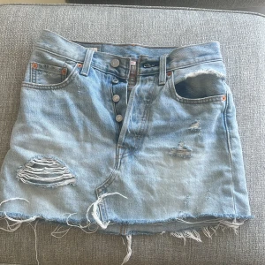 Ljusblå jeanskjol från Levi's - Säljer en ljusblå jeanskjol från Levi's med slitna detaljer, fransig nederkant och klassiska fickor både fram och bak. Kjolen har knappar framtill och en avslappnad, cool vibe. Perfekt för dig som gillar en edgy streetstil.