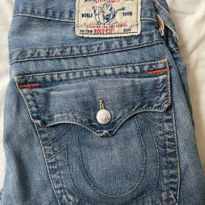 True Religion Bootcut Jeans Blå - Säljer ett par klassiska blå bootcut jeans från True Religion med ikoniska bakfickor och silverfärgade knappar. Jeansen har orange detaljer vid fickorna och en snygg tvättad finish. Perfekta för dig som gillar en avslappnad men ändå trendig look.