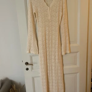 Lång stickad spetsklänning BYIC - Superfin långklänning från BYIC i ljusbeige stickat material med spetsmönster. Klänningen har v-ringning, knappar framtill och långa ärmar med lätt utsvängd avslutning. Perfekt för sommarens alla tillfällen!