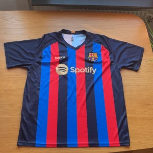 FC Barcelona Lewandowski matchtröja XL - Säljer en officiell FC Barcelona matchtröja i storlek XL med Lewandowski och nummer 9 på ryggen. Tröjan har klassiska blå och röda ränder, klubbmärke på bröstet och Spotify som sponsor. Tillverkad i lätt och ventilerande polyester, perfekt för fotboll eller att bära som supporter.