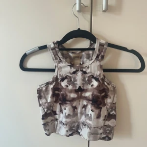 Träningstopp från Stronger - Cool croppad linnetopp från Stronger med unikt mönster i bruna och beige toner. Toppen har sportig passform med breda axelband och ärmlös design, perfekt för träning eller chill dagar. Materialet är stretchigt och andas, så du kan röra dig fritt.