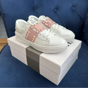 Valentino Rockstud sneakers vit/rosa - Snygga Valentino Rockstud sneakers i vitt skinn med ljusrosa band och ikoniska nitar. Klassisk rund tå, platt sula och vita snören. Perfekta för dig som vill ha en lyxig och trendig look med en touch av rosa detaljer.