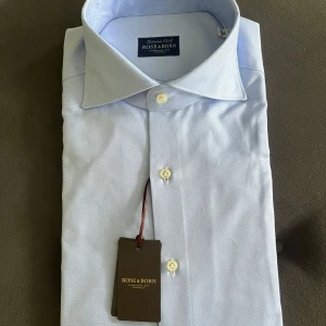 Oanvänd Ljusblå skjorta från Rose & Born - Helt oanvänd med taggar! Klassisk ljusblå skjorta från Rose & Born med snygg krage och vita knappar. Skjortan är tillverkad i bomull och har en stilren, tidlös design som passar perfekt till både jeans och kostym. Perfekt för dig som gillar en clean och fräsch look.