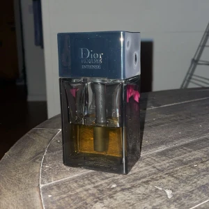 Dior Homme Intense Eau de Parfum - Dior Homme Intense Eau de Parfum i en stilren, fyrkantig glasflaska med mörkgrått lock och silverfärgad Dior-logga på toppen. Flaskan har en transparent design som visar den bärnstensfärgade vätskan inuti. En klassisk och lyxig herrdoft.