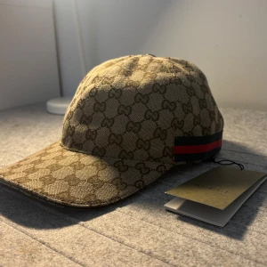 Beige Gucci keps - Skicka dm för video. Storlek ”M”. Snygg beige keps från Gucci med klassiskt GG-monogram över hela kepsen. Kepsen har justerbart spänne bak i brunt skinn och ikoniska röd-gröna ränder på sidan. Tillverkad i bomullscanvas med exklusiv känsla, perfekt för dig som vill sticka ut.