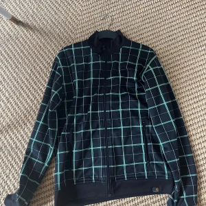 Rutig svart Carhartt Grid jacka L - Svart jacka från Carhartt i storlek L men passar som M med rutigt mönster i vitt och grönt. Jackan har dragkedja framtill, ståkrage och två fickor med dragkedja. Tillverkad i 100% polyester och har en sportig, clean vibe som passar perfekt till streetwear.