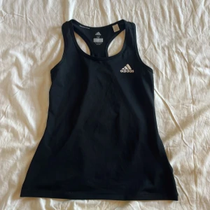 Svart träningstopp Adidas Climalite - Svart sportig linnetopp från Adidas med klassisk logga framtill och tre vita ränder på ryggen. Modellen är ärmlös med brottarrygg och tillverkad i lätt och ventilerande syntetmaterial, perfekt för träning och aktiva dagar.