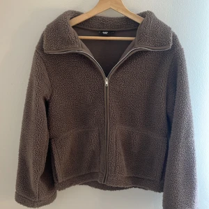 Brun teddyjacka från 157, stl S - Mysig brun teddyjacka från 157 i storlek S. Jackan har en hög krage, hel dragkedja framtill och två stora fickor. Perfekt för kyliga dagar när du vill vara både varm och snygg. Den fluffiga teddytexturen ger en chill och trendig vibe.