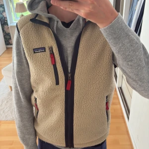 Beige fleeceväst från Patagonia S - Säljer en beige fleeceväst från Patagonia med marinblå detaljer och röda dragkedjor. Västen har hög krage, tre fickor med dragkedja och klassisk Patagonia-logga på bröstet. Perfekt att slänga över en hoodie för extra värme.
