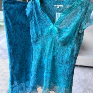 Turkos/blå mönstrad set kjol och blus  - Snygg turkos/blå kjol och blus från XLNT med svart paisleymönster och blommor. Blusen har v-ringning, korta ärmar och lätt utsvängd nederdel. Kjolen har samma mönster och nederdel, samt fin  broderad detalj vid midjan. Tunt och luftigt material, perfekt för varma dagar. Superfin färg som verkligen poppar! (Står strl 42 men den passar from 38)