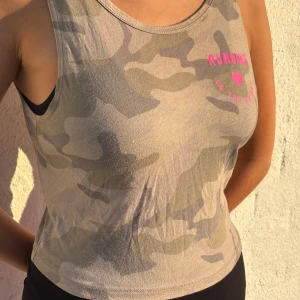 Camouflage croppad linnetopp - Säljer en croppad linnetopp i camouflage-mönster med grå och beige toner. Toppen har ärmlös design, rund halsringning fram och djup v-ringning i ryggen. På bröstet finns en rosa text med hjärta där det står 'RUNNING TO THE FUTURE'. Perfekt för dig som gillar sportig och edgy stil.