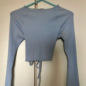 Blå croppad topp med snörning NA-KD - Trendig ljusblå croppad topp från NA-KD med långa ärmar och ribbad struktur. Toppen har öppen rygg med snygg snörning och volangkant vid hals och nederdel. Perfekt för dig som vill sticka ut med en cool och modern look.