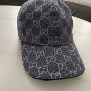 Blå Gucci keps - Snygg blå keps från Gucci med ikoniskt GG-mönster över hela kepsen. Justerbar rem baktill i blått och diskret guldfärgad GG-detalj på sidan. Tillverkad i bomullsmaterial med vit insida och klassisk böjd skärm. Perfekt för dig som vill sticka ut med lyxig streetstyle.