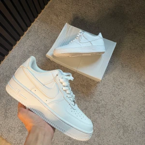 Nike Air Force 1 vita - Klassiska Nike Air Force 1 sneakers i helvitt skinn med lågt skaft och perforerad tå. Snygg och clean design med vit sula, snörning och ikonisk Nike-logga på sidan och hälen. Perfekta för dig som vill ha en tidlös och fräsch look. Storlek 40 endast använda en gång.