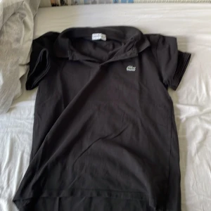 Svart pikétröja från Lacoste - Klassisk svart pikétröja från Lacoste i storlek S. Tröjan har korta ärmar, krage och den ikoniska gröna krokodilloggan på bröstet. Tillverkad i bomull för en skön och luftig känsla. Perfekt för en clean och sportig stil. Använd många gånger men är fortfarande i bra skick. Kan göra snabb affär.