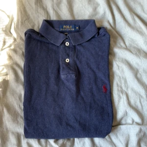 Marinblå pikétröja Polo Ralph Lauren - Snygg marinblå pikétröja från Polo Ralph Lauren i slim fit-modell. Klassisk krage, tre knappar och röd broderad logga på bröstet. Kortärmad och tillverkad i mjuk bomull, perfekt för en clean och stilren look. Passar även en M/L och en S/M.  Nypris ca 1500.