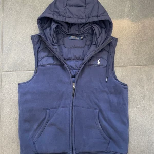Marinblå Ralph Lauren väst  - Mörkblå väst med huva från Polo Ralph Lauren. Västen har dragkedja, två fickor fram och ribbade muddar med vita ränder.                                       Storlek:M.                                                                          Skick: Mycket bra😊