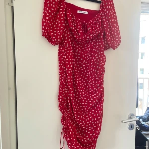 Röd blommig klänning från Adoore - Superfin röd klänning från Adoore med vita blommor, puffärmar och snörning framtill. Klänningen har omlottkänsla, rynkade detaljer längs sidorna och är i en lätt, flowy kvalitet. Perfekt för sommarens alla tillfällen! Passar en medium eftersom Adoore är små i storleken.