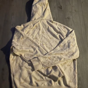 Beige hoodie från Lacoste LIVE - Säljer en beige hoodie från Lacoste LIVE i unisexmodell. Hoodien har all-over print med Lacoste-loggor och krokodiler i ljusbeige, snörning i huvan och en klassisk känguruficka framtill. Mjuk bomullskänsla och ribbade muddar vid ärmslut och nederkant.