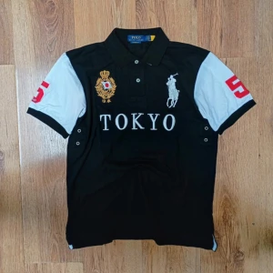 Polo Ralph Lauren Piké - Tokyo - Säljer en snygg pikétröja från Polo Ralph Lauren med svart kropp, vita ärmar och röda siffror. Stora broderier framtill med Tokyo-text, guldemblem och klassisk polospelare. Krage och knappar framtill. Perfekt för dig som gillar sportig och stilren look.