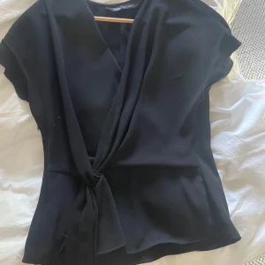 Svart omlottopp från Zara - Svart omlottopp från Zara med knyt framtill. Toppens material är mjukt och har en lätt stretch. Den har korta ärmar och en snygg v-ringning. Den har en liten defekt med att en som har gått upp så de syns men de är bara vika in eller sy in, super enkelt 