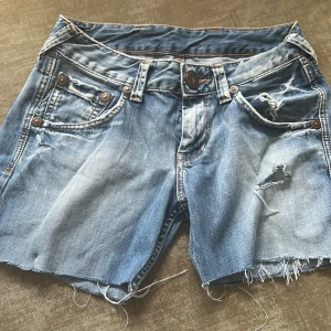 Blå jeansshorts från Pepe Jeans - Snygga blå jeansshorts (från början jeans) från Pepe Jeans med slitna detaljer, råa kanter och trasiga partier. Klassisk femficksmodell med knappar och dragkedja. Baksidan har en cool detalj med rosa söm på ena fickan. Shortsen var från början ett par jeans som sedan gjort om till shorts. Size: 28/32 och rekommenderas tvättas i 40 grader. Perfekta för dig som vill ha ett par snygga shorts nu i sommar!!
