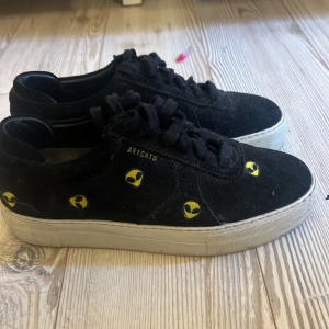 Svarta sneakers från Axel Arigato - Svarta sneakers från Axel Arigato med coola gula alien-huvuden broderade på sidorna. Skorna har vit platt sula, snörning och är gjorda i mocka.