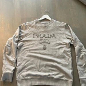 Prada Grå sweatshirt  - Snygg grå sweatshirt från Prada Milano med upphöjt logotyptryck framtill. Tröjan har rund hals, ribbade muddar och är tillverkad i mjuk bomull. Perfekt för dig som gillar stilrena designer och exklusiva märken.