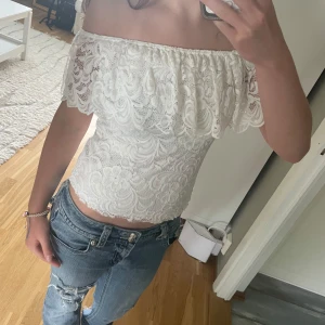 Vit offshoulder spets topp - Superfin vit offshoulder topp i spets med volang upptill. Sitter tight och har en croppad passform som framhäver midjan. Perfekt till jeans eller kjol för en romantisk look. Spetsen har ett vackert mönster och toppen är ärmlös med axelbandslös design.