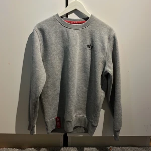Sweatshirt grå Alpha Industries - Snygg grå sweatshirt från Alpha Industries med liten svart logga på bröstet och röd detalj vid nederkanten. Klassisk passform, rund hals och långa ärmar. Perfekt för dig som gillar stilren streetwear och vill ha något bekvämt till vardags.