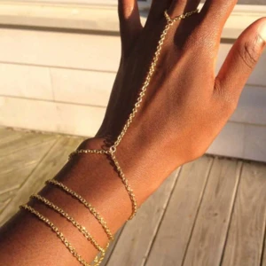Guldigt handkedje-armband - Snyggt och trendigt handkedje-armband i guldtonad metall. Kedjan går från handleden och fäster runt ett finger, vilket ger en unik och stilren look. Perfekt accessoar för att lyfta din outfit med lite extra edge.