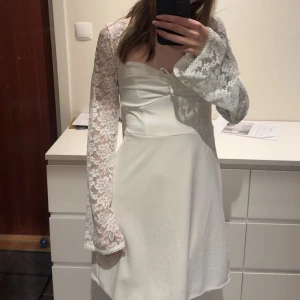 Vit spetsklänning  - Superfin vit klänning med långa, blommiga spetsärmar och öppen rygg med knyt. Använd en gång så verkligen i nyskick! Kostar 699 ny