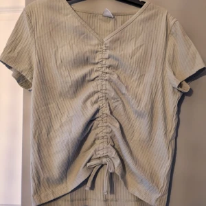 Grön ribbad topp med snörning - Snygg grön ribbad topp med kort ärm och v-ringning. Toppen har en cool snörningsdetalj framtill som ger en trendig look och gör att du kan justera passformen. Perfekt till jeans eller kjol för en avslappnad stil.