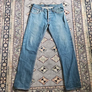 Nudie Jeans blå straight fit jeans - Säljer ett par klassiska blå jeans från Nudie Jeans i rak modell. Jeansen har fem fickor, orangea sömmar och Nudie-logga på bakfickan. Tillverkade i 100% bomull med normal passform och snygg tvättad look. Hör av er vid minsta fundering🙏