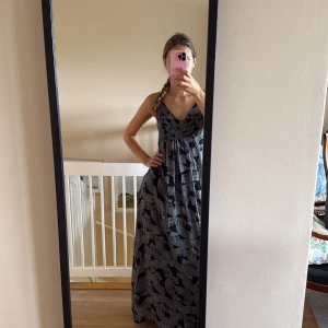 Svart och grå långklänning med mönster - Långklänning med svart och grått abstrakt mönster, omlott-v-ringning och smala axelband. Klänningen har en luftig passform och faller snyggt ner till golvet. Perfekt för dig som gillar en stilren men ändå unik look.