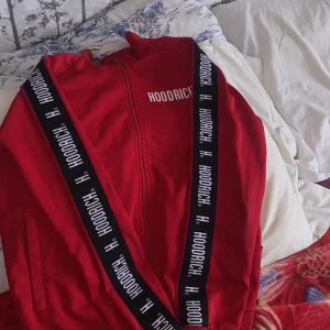Röd Hoodrich track jacket XS - Röd track jacket från Hoodrich med dragkedja framtill och svarta band med vit logga längs ärmarna. Jackan har ståkrage, fickor på sidorna och sportig vibe. Perfekt för dig som gillar streetwear och vill sticka ut med färg.