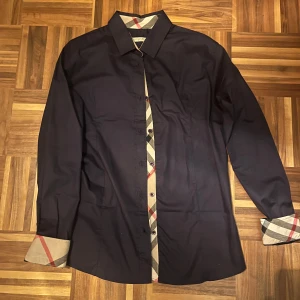 Mörkblå Burberry skjorta - Snygg mörkblå skjorta från Burberry Brit med klassiska rutiga detaljer på insidan av kragen, knappslån och ärmsluten. Storleksettiketten har blivit avklippt men passar som en small eller en slim fit medium. Annars i mycket fint skick utan några tecken på skavanker.