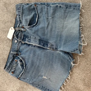 Levi's 501 jeansshorts med rå kant - Klassiska blå jeansshorts från Levi's 501 med rå, fransig kant och femficksmodell. De har en avslappnad passform och ikoniska detaljer som synliga nitar och bakfickor med signatursöm. Hög i midja.