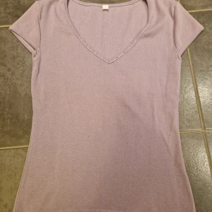 Ljuslila ribbad topp med v-ringning - Ljuslila topp med korta ärmar och v-ringning. Ribbad struktur och håldetaljer längs ringningen, där även band kan dras om man vill dra ihop urringningen (ingår dock inte)