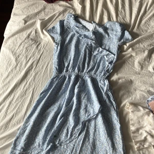 Blommig blå kortklänning Hampton Republic - Supersöt blå kortklänning från Hampton Republic med små vita blommor, volangdetaljer och omlottkänsla upptill. Klänningen har korta ärmar och resår i midjan för en skön passform. Perfekt för varma dagar och somriga vibes!