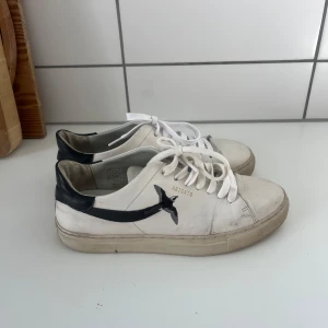 Vit-svarta sneakers från Axel Arigato - Snygga vita sneakers från Axel Arigato med svart fågelmotiv på sidan och svart hälparti. Klassisk låg modell med rund tå, vita snören och platt sula. Skorna är i skinn och har en stilren, minimalistisk design med diskret guldtext på sidan.