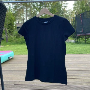 Svart ribbad topp med kort ärm - En svart ribbad topp med rund halsringning och korta ärmar. Toppen har en enkel och clean look med vertikala ribbade ränder som ger lite extra struktur. Perfekt att matcha med jeans eller kjol för en stilren vibe.