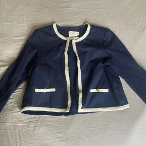 Mörkblå kavaj med vita detaljer - Snygg mörkblå kavaj från Monet Le Martre med vita, strukturerade kanter och dekorativa fickor framtill. Jackan har rund halsringning, lång ärm och guldiga knappar på fickorna. Perfekt för dig som gillar klassisk men trendig stil.