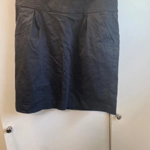 Svart kortkjol från Lindex - En svart, klassisk kortkjol från Lindex i storlek 38. Kjolen har en enkel och stilren design med diskreta veck framtill och dragkedja bak. Perfekt att matcha med en snygg topp eller stickad tröja för en clean look.