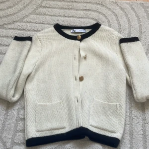 Vit kofta med svarta detaljer från Zara - Säljer en stilren vit kofta från Zara med svarta kanter vid hals, ärmslut och nederkant. Koftan har guldknappar framtill, två fickor och är stickad i ett mjukt material. Perfekt för lager-på-lager och snygg till jeans eller kjol. 