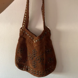Brun mocka axelväska med nitar - Säljer en brun axelväska i mocka från American Eagle Outfitters. Väskan har nitar och öglor i mönster på framsidan och längs axelremmen. Mjuk form och stängs med magnetknapp. 