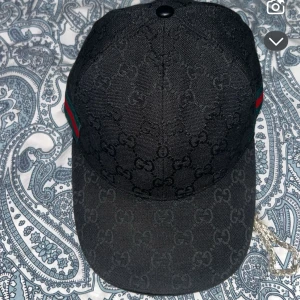 Svart keps från Gucci med GG-mönster - Snygg svart keps från Gucci med klassiskt GG-monogram över hela kepsen. Justerbar rem baktill i läder och diskreta gröna och röda detaljer på sidan. Tillverkad i bomull och läder för en exklusiv känsla.
